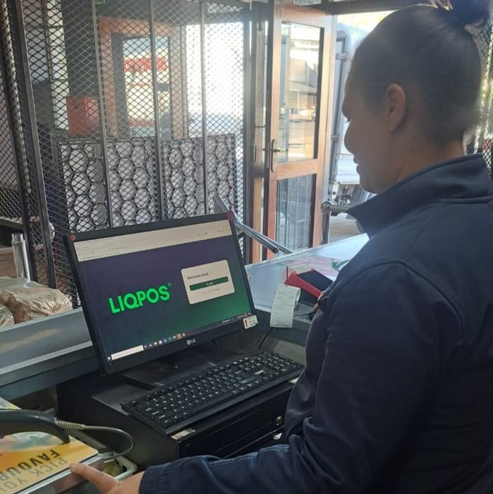 Maderé, cashier at The Grapevine Blue Bottle Liquors, standing at the till counter beside a monitor showing the LIQPOS login screen.