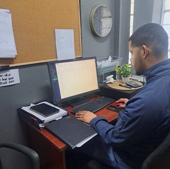 Vicus, manager at The Grapevine Blue Bottle Liquors, seated at a back office desk working on a computer monitor that shows a LIQPOS stock list.